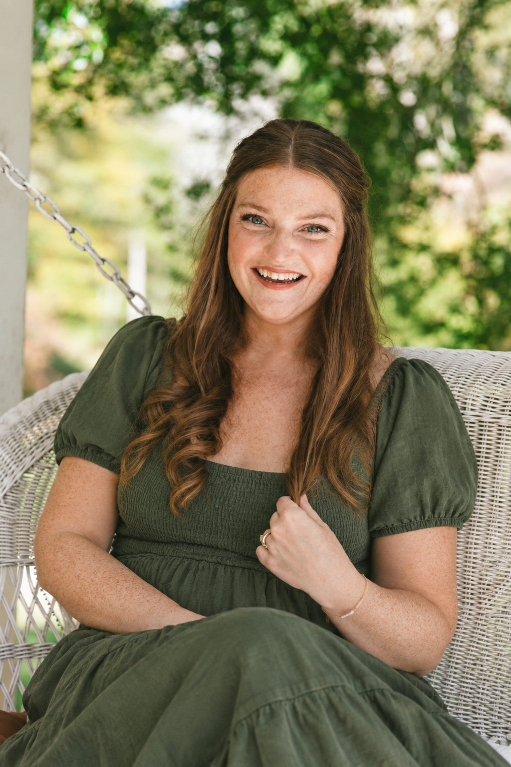 title Mara, founder of Red Finch, sitting on a porch swing in a green dress, smiling with trees in the background.